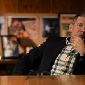 A man sitting behind a desk, leaning back in his chair and scratching his chin, mid-conversation.