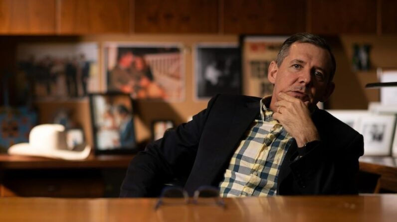 A man sitting behind a desk, leaning back in his chair and scratching his chin, mid-conversation.